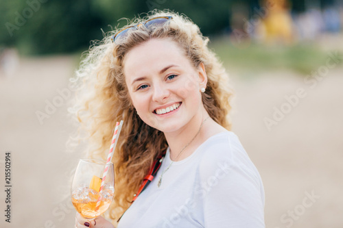 Fotografie, Obraz Close up portrait of beautiful European female with light wavy hair, toothy smile, holds cold fresh beverage, enjoys sunny good weather, poses against blurred beach background