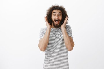 Wall Mural - Portrait of happy overwhelmed handsome male model with beard and curly hair, screaming from surprised and positive emotions, holding palms near face, staring at camera joyfully over gray wall