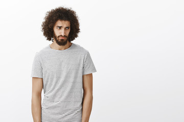 Wall Mural - Indoor shot of handsome gloomy hispanic guy in striped t-shirt, looking from under forehead right with miserable jealous expression, feeling disappointemnt after losing something over gray background