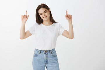 Beautiful healthy and stylish female student in jeans and t-shirt, raising hands high and pointing up with index fingers, smiling joyfully, giving direction or showing great place for advertisement