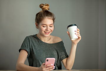 Young attractive woman with blonde hair knot, typing text message on smart phone at home over grey wall. Studio image of young woman sitting at a table with a take-away coffee holding mobile phone.