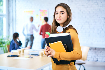 female student standing in classroom holding books. portrait of young woman looking at camera indoor