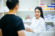© twinsterphoto - Asian female pharmacist doctor in professional gown explaining and giving advice with male client in drugstore shop.