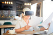 © pressmaster - Modern senior financier with papers watching virtual presentation in vr goggles while sitting in cafe