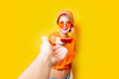 © Masson - Young style girl with orange glasses give to you her phone on yellow background. Clothes in 1980s style