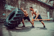 © zamuruev - Two muscular athletes training. Muscular fitness shirtless man moving large tire other motivate him and hold big hummer in street gym. Concept lifting, workout training.
