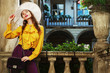 Outdoor fashion portrait of young beautiful woman wearing stylish white hat, yellow blouse with frills, holding violet velvet bag, posing in street, near old european architecture. Copy, epmty space