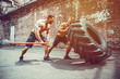 © zamuruev - Two muscular athletes training. Muscular fitness shirtless man moving large tire other motivate him and hold big hummer in street gym. Concept lifting, workout training.