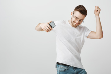 Wall Mural - Joyful pleased and carefree exciting handsome unshaven guy in glasses holding black smartphone dancing sliding on floor while listening music happily in wireless earphones over gray background