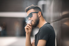 A Young Handsome White Bearded Man In Black T-shirt Smokes A Cigarette In The Street In The Spring. Close Up.