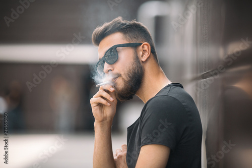Fotomural A young handsome white bearded man in black t-shirt smokes a cigarette in the street in the spring