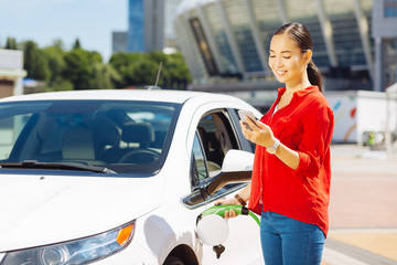 Wall Mural - Modern age. Confident young woman looking at her smartphone while holding a fuel nozzle