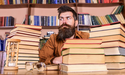 Leinwandbilder - Bookworm concept. Man on strict face sit between piles of books, while studying in library, bookshelves on background. Teacher or student with beard sit at table with hourglass and glasses, defocused.