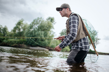 Fisherman in action. He stands in water and holds fly rod in one hand and spoon in the other one. Also adult has a fishing net on the back. Green trees are at the edge of water.