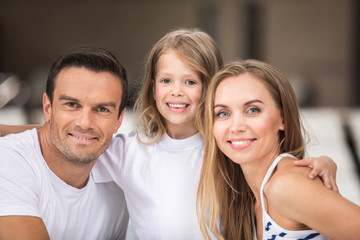 Wall Mural - Happy together. Waist up portrait of smiling parents bonding to their kid. Small child is standing and embracing mother and father with content while they are sitting and looking straight with delight