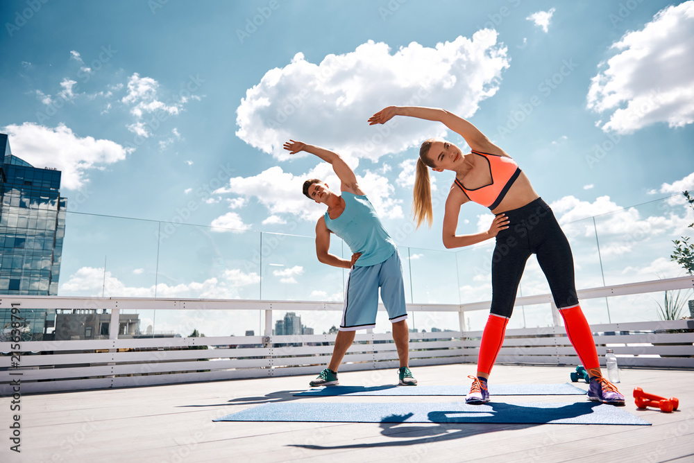 Positive couple are exercising jointly on terrace of high city building ...