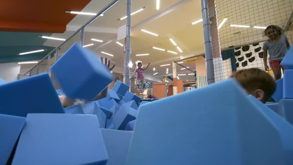 Wall Mural - Low angle view of two children, boy and girl, jumping into cube pit and having play fight with blue soft foam cubes while two other girls bouncing on trampoline in the background