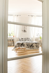 Wall Mural - View through a glass and wood door to a spacious white bedroom interior with a bed with linen standing on a herringbone parquet floor. Real photo.