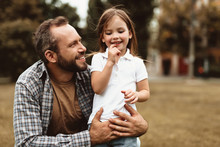 Side View Of Delighted Man Squatting Near Small Kid And Holding Her. Little Girl Is Standing With Smile And Enjoying Leisure Outdoors
