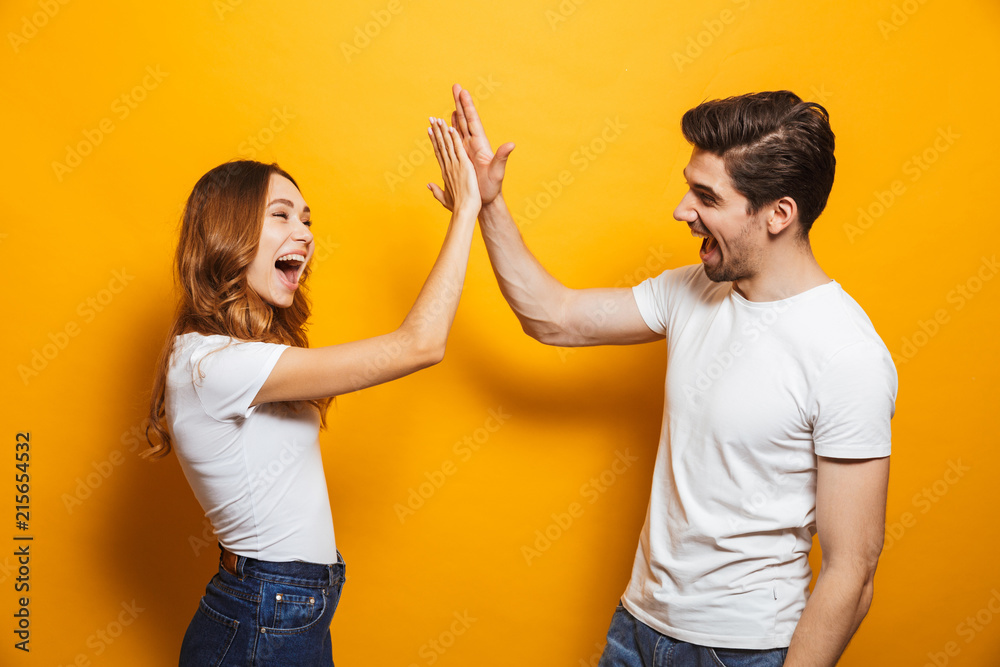 Image of friendly young people man and woman in basic clothing laughing and giving high five, isolated over yellow background - obrazy, fototapety, plakaty 