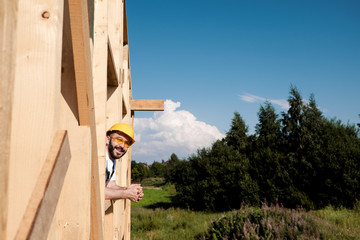 The man builder sits on the edge of the roof of the frame house, in a yellow helmet and gray overalls and a white T-shirt. Looks from the window of construction.