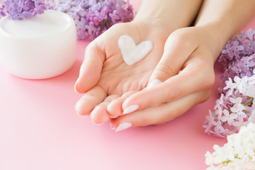 Wall Mural - Young, perfect, clean woman's hands. White jar of natural herbal skin cream on pastel pink background. Heart shape created from cream. Love a body. Beautiful branches of lilac blossoms. Fresh flowers.