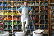 Full length portrait of modern artisan wearing apron standing on step ladder and posing in workshop against shelves with materials, copy space