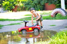 Cute 2 Year Old Boy In A White Shirt Playing With A Red Car Near A Puddle In The Park