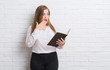 © Krakenimages.com - Young adult woman standing over white brick wall reading a book cover mouth with hand shocked with shame for mistake, expression of fear, scared in silence, secret concept