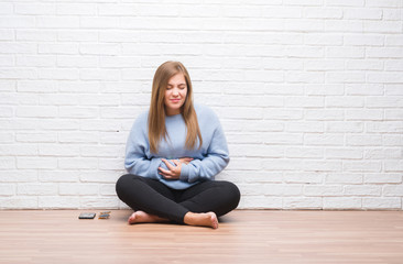 Canvas Print - Young adult woman sitting on the floor in autumn over white brick wall with hand on stomach because indigestion, painful illness feeling unwell. Ache concept.