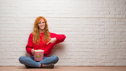 Canvas Print - Young redhead woman sitting over brick wall eating popcorn very happy pointing with hand and finger