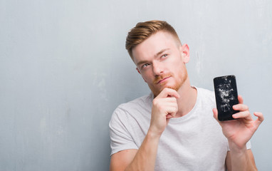 Wall Mural - Young redhead man over grey grunge wall holding broken smartphone serious face thinking about question, very confused idea