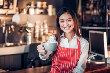 Wall Mural - asian woman barista wear red apron holding hot coffee cup and smiling at bar counter with happy emotion,Cafe restaurant service concept,selective focus on smiling face.