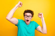 Handsome curly-haired shocked young guy wearing casual green t-shirt and glasses, showing surprised gesture. Isolated over yellow background
