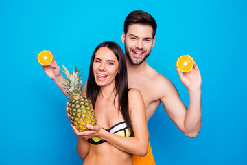 Young brunette lady laugh and hold pineapple, winks and make funny face near the man with beard wearing in swim suit isolated on blue background