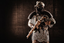Post Apocalyptic Soldier In Black Mask And Glasses, Wool Field Cap And Handmade Armor From Car Tires And Hauberk, Standing At Attention With Submachine Gun On Shoulder, Black Background Studio Shoot