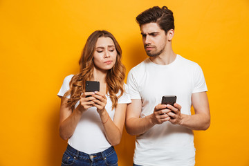 Wall Mural - Image of curious man and woman frowning and peeking at each others cell phones, isolated over yellow background