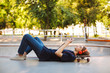 Young skater in orange headphones lying on skateboard thoughtfully using cellphone with skatepark on background