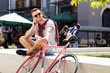 © proimagecontent - Handsome young hipster man in sunglasses sitting near the bicycle, smiling and speaking on the smartphone in the sunny street