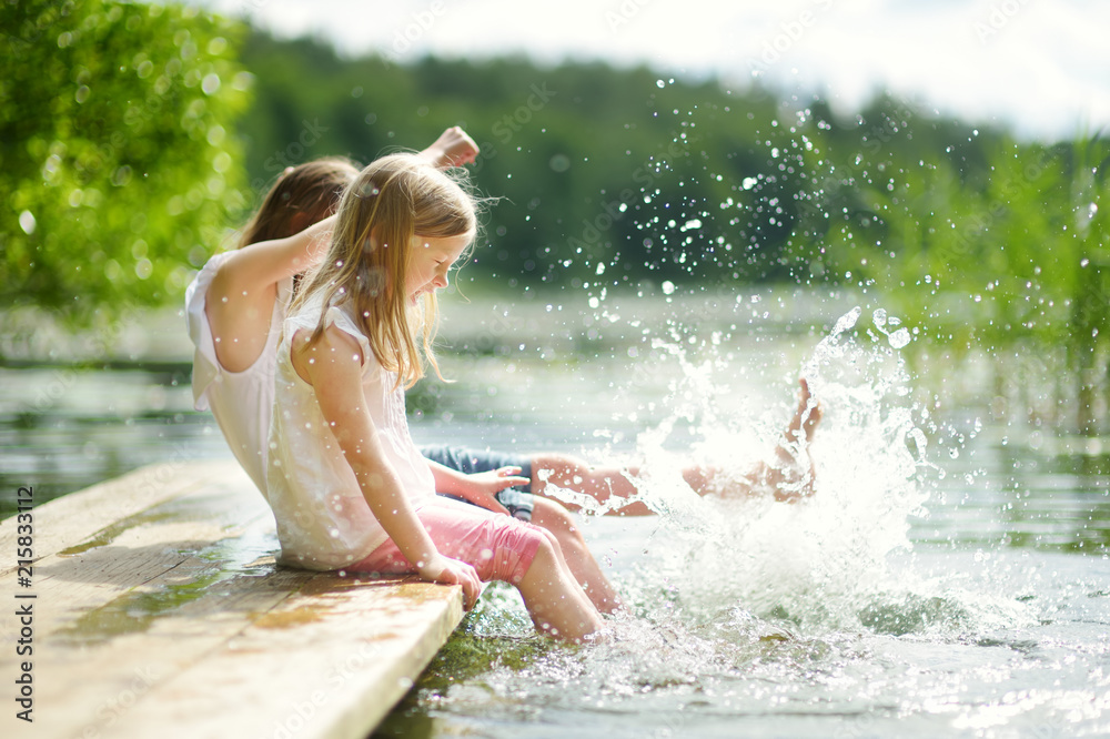 Two cute little girls sitting on a wooden platform by the river or lake dipping their feet in the water on warm summer day - obrazy, fototapety, plakaty 