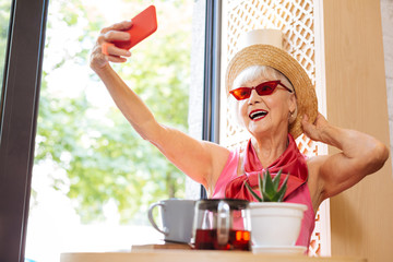 Beautiful accessory. Cheerful positive woman touching her hat while taking a selfie