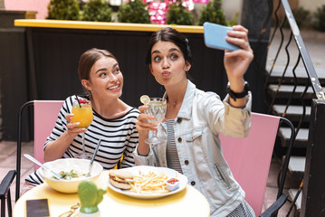 Wall Mural - Funny faces. Two beaming dark-haired women making funny faces while making photo having lunch