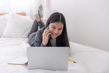 Poster - Asian woman laying on bed talking on mobile phone while browsing data on laptop in bedroom.work at home.e-larning concept.study online.