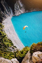 Myrtos Beach, Kefalonia Island, Greece. Figure Of A Parachutist Skydiver With Orange Parachute Against A Blue Lagoon Bay Beach