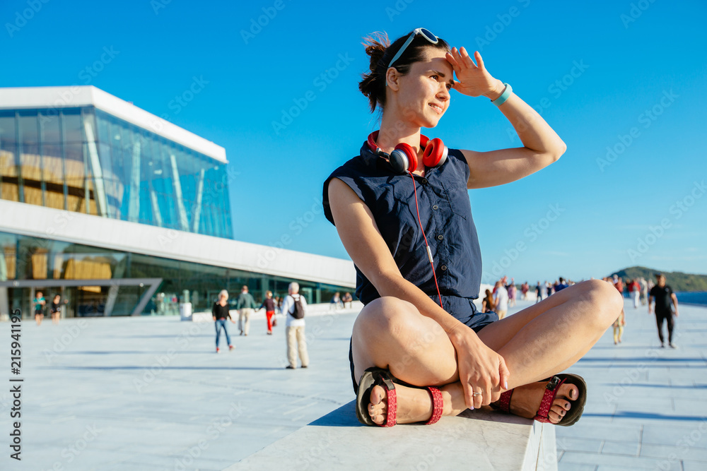 Obraz na płótnie Young hipster woman traveler with red headphones resting on border after walking in Oslo city over Opera House urban background, happy, smiling, looking in sunset. w salonie