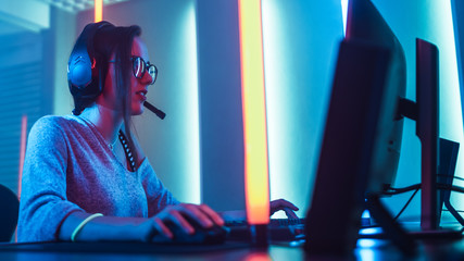 Wall Mural - Low Angle Shot of the Beautiful Friendly Pro Gamer Girl Playing in Online Video Game and Streaming it, Wearing Headset Talks with Her Fans and Team into Microphone. Background Cool Neon Retro Colors.