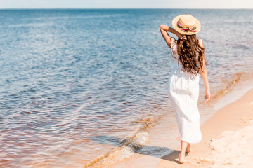 Wall Mural - back view of woman in straw hat and white dress walking on beach near sea