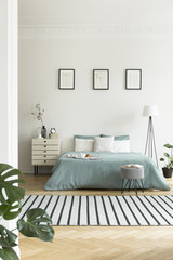 Real photo of bright bedroom interior with three simple posters, striped carpet and pastel green sheets on double bed