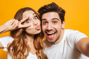 Sticker - Portrait of caucasian man and woman taking selfie photo and showing peace sign, isolated over yellow background