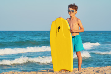 A happy boy in sunglasses in swimming trunks stands on the beach on golden sand and holds a swimming board, boogie board of yellow color against the blue sky and the sea with small waves in sunny sun